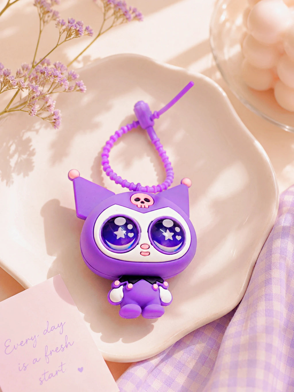 A cute purple cartoon keychain charm with large starry eyes and a coiled purple strap, styled on a white scalloped dish with dried flowers and a lavender check cloth.