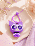 A cute purple cartoon keychain charm with large starry eyes and a coiled purple strap, styled on a white scalloped dish with dried flowers and a lavender check cloth.