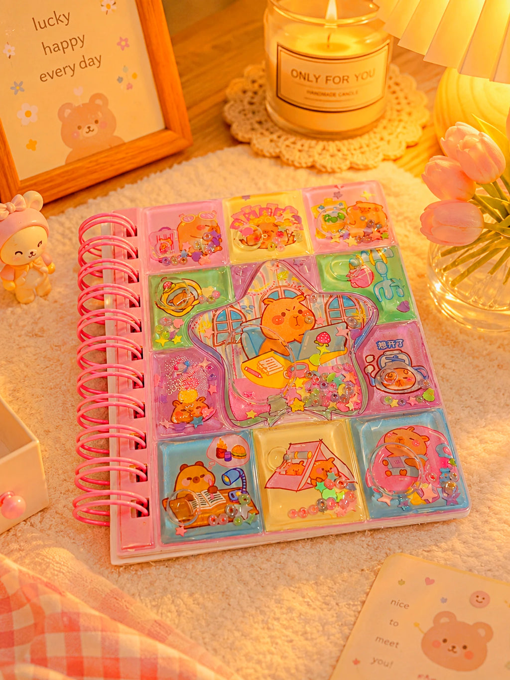 A pastel-colored spiral-bound notebook with cartoon animal illustrations and a pink spiral binding, styled on a soft cream fabric with a candle, flowers, and a small plush toy nearby.
