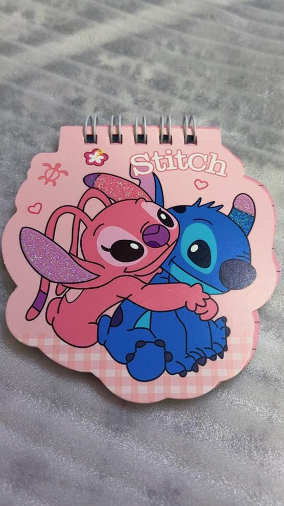 A small spiral-bound notebook featuring a pink and blue cartoon character design with the word 'Stitch' on the cover, placed on a gray textured fabric surface.
