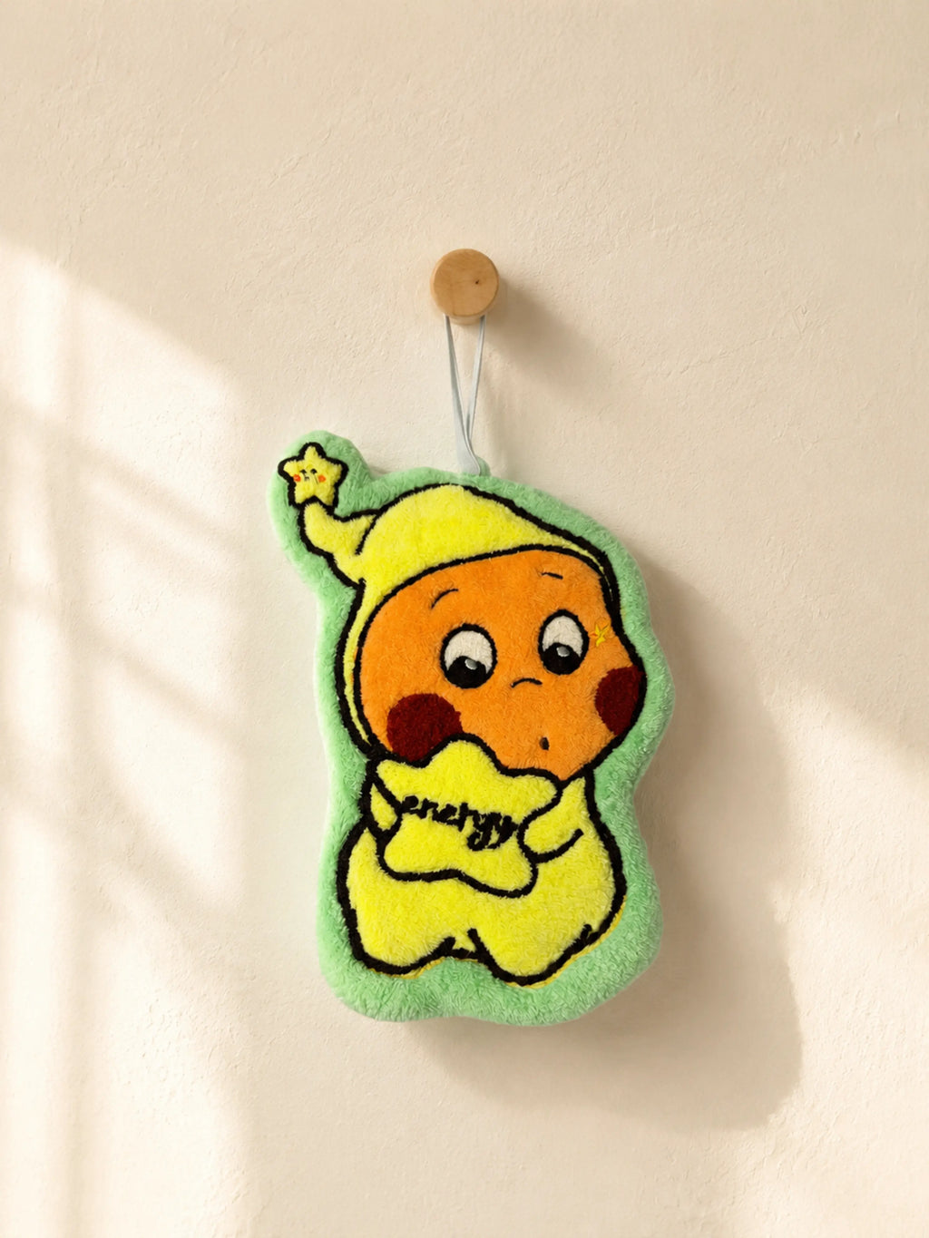 Cute mint green character holding a yellow star hand towel hanging from a wooden wall peg in a warm minimalist bathroom